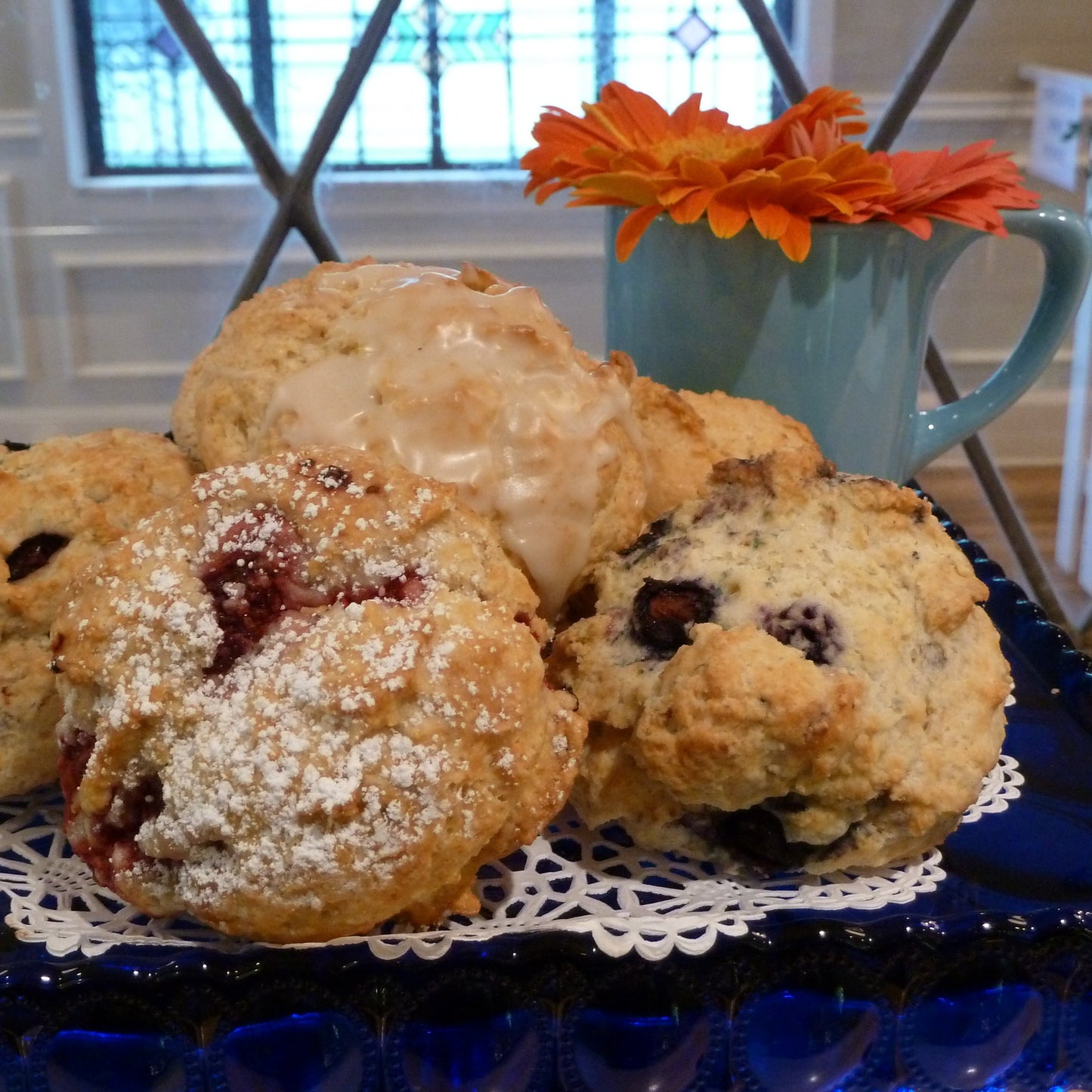 Fresh scones on a blue glass plate with lace doily, berry and glazed varieties, flowers in background.
