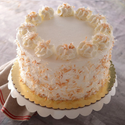 Round cake with white frosting, toasted coconut topping and sides, on gold board and scalloped white stand.
