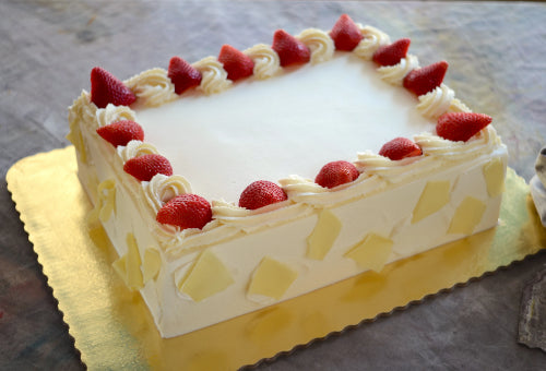 Rectangular cake with white frosting, cream swirls, strawberries, white chocolate shards, on gold base.
