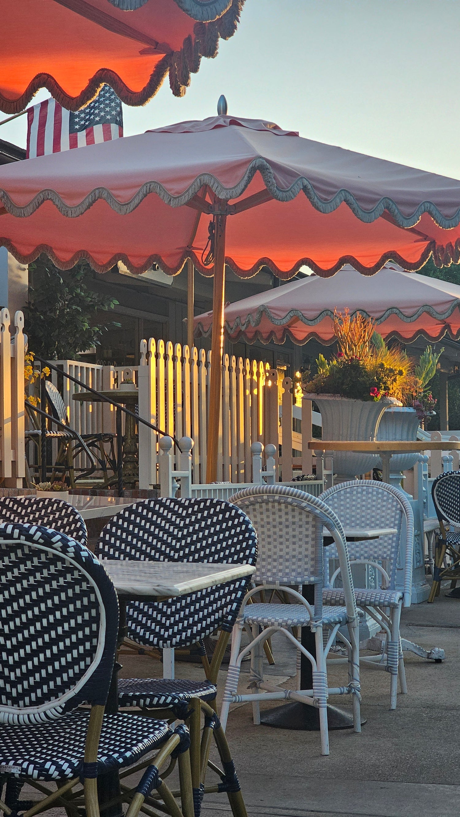 Outdoor café with red umbrellas, woven chairs, and picket fence. Sunlight adds warmth to a charming setup.