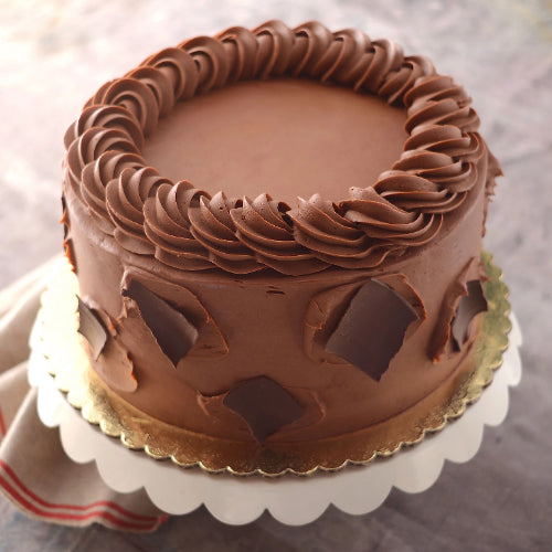 Round cake with smooth chocolate frosting, swirl border, dark chocolate shards, on gold board and stand.a