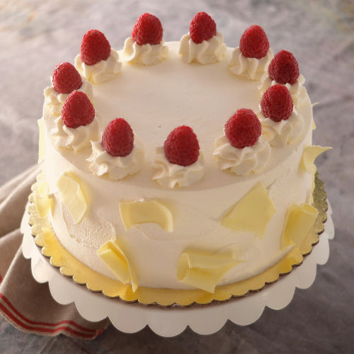Round cake with white frosting, raspberries on whipped cream, white chocolate curls, on gold board and stand.