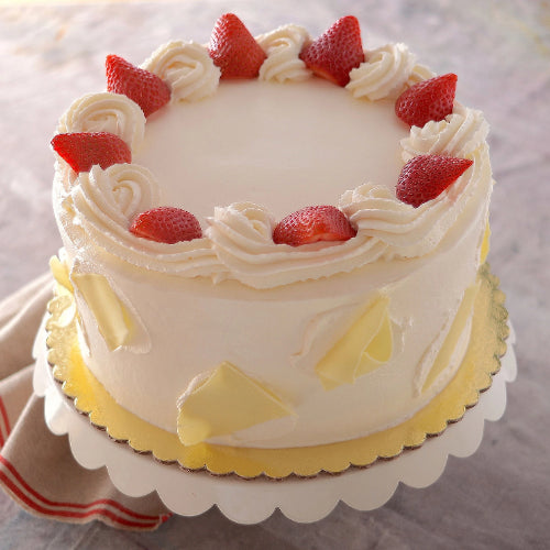 Round cake with white frosting, whipped cream swirls, strawberries, and white chocolate shards on gold board.