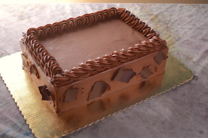 A rectangular cake covered in smooth chocolate frosting with a piped chocolate border around the top edge. The sides are decorated with large dark chocolate shards, and the cake is displayed on a gold cardboard base.